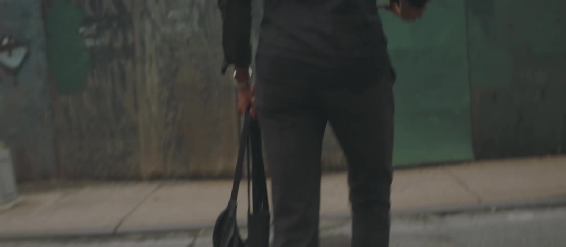 A black male walks across a city street and down a sidewalk while listening to his smartphone