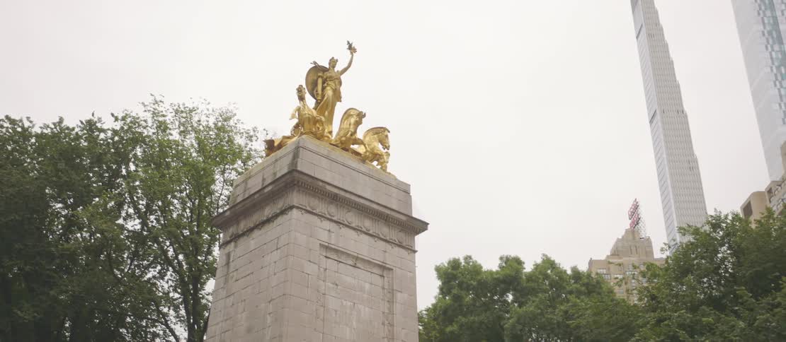 A monument with golden statues on top of it sits near trees in a city