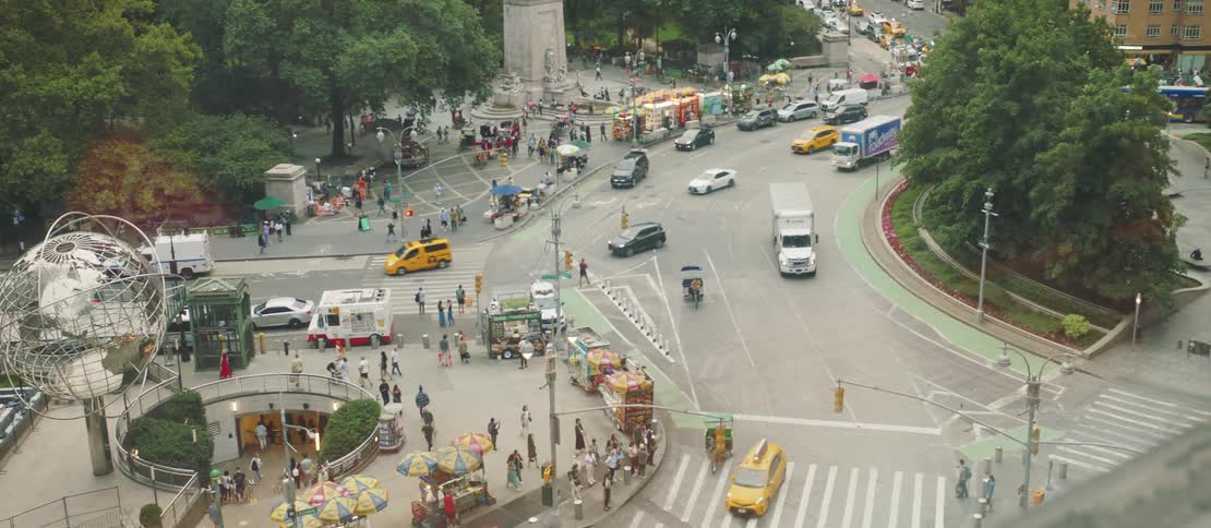 An aerial view of a bustling urban plaza filled with pedestrians food carts and vehicles including yellow taxis and delivery trucks in the foreground a large metallic globe sculpture stands prominently next to a subway entrance while the scene
