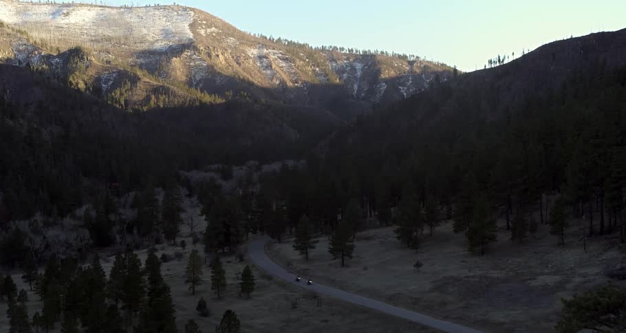 A landscape gave an asphalt road with two motorcycles passing and there is a pine forest around and in the background there is a big mountain