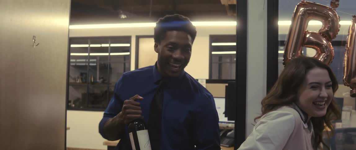 Two people happily walk into a room with birthday balloons on the wall and one of the people is holding a bottle of drink