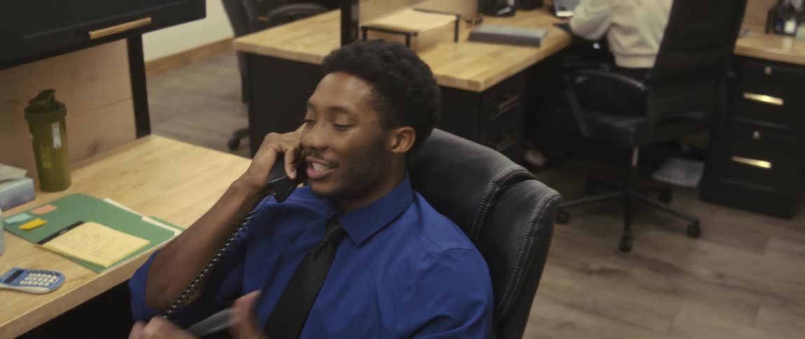 A man is sitting on a chair in an office and is talking on the phone while he is gesturing and he is happy