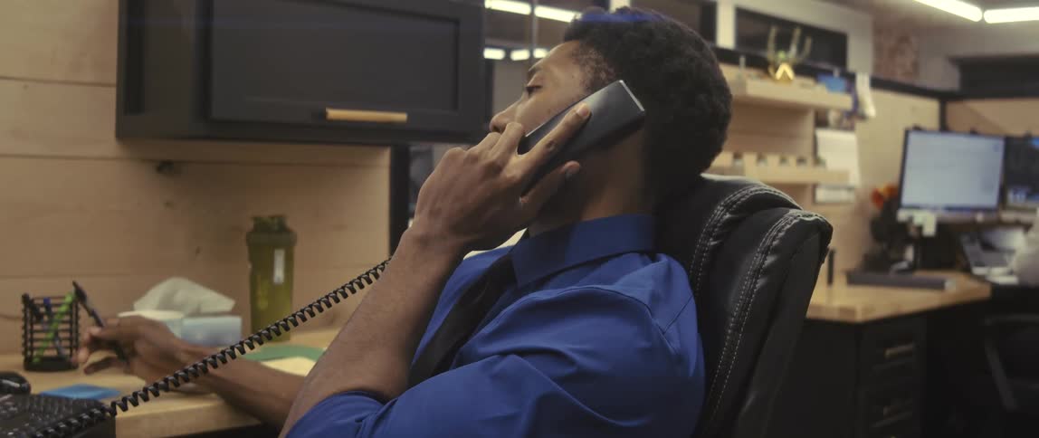 A relaxed office worker wearing a blue shirt talking on the phone on his cubicle