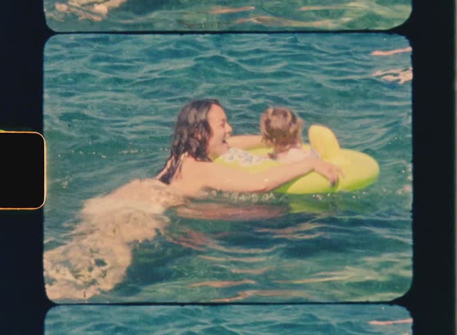 Super 8 film of a mother swimming in the water guiding a small child who is floating in an inflatable ring possibly enjoying a relaxed moment together in a natural or outdoor setting