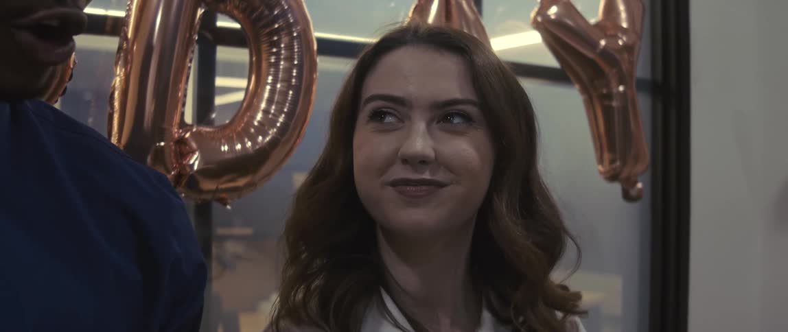A young woman at an office birthday party smiles and nods as someone beside her speaks