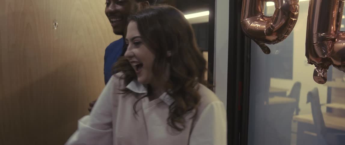 A man and a woman walk into a room that has balloons in the window and they are happy and talking while they are looking straight ahead