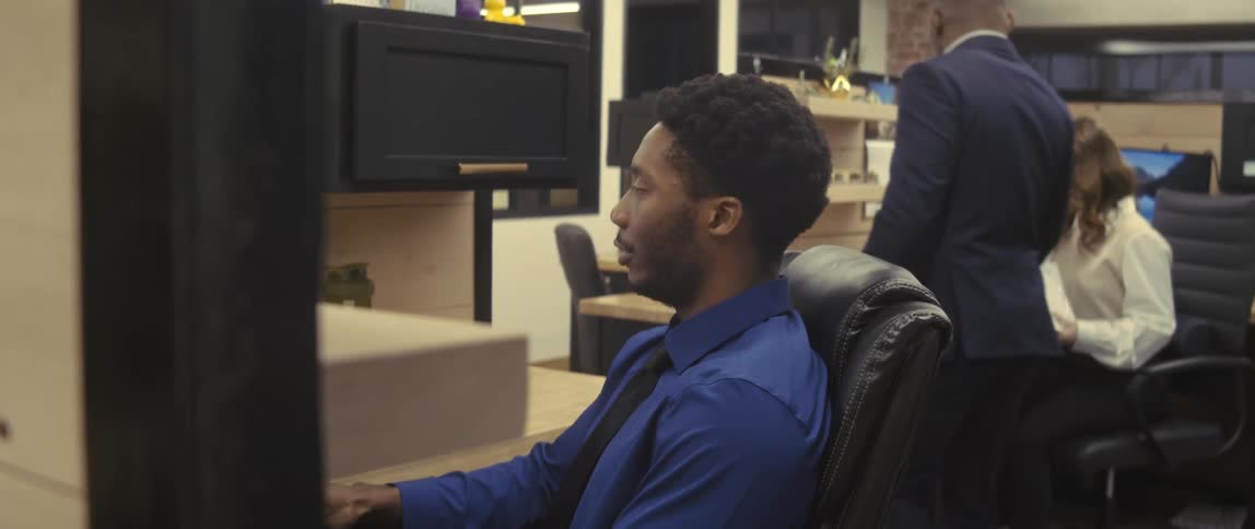 Two people in an office environment sitting working in front of the computer and one man standing