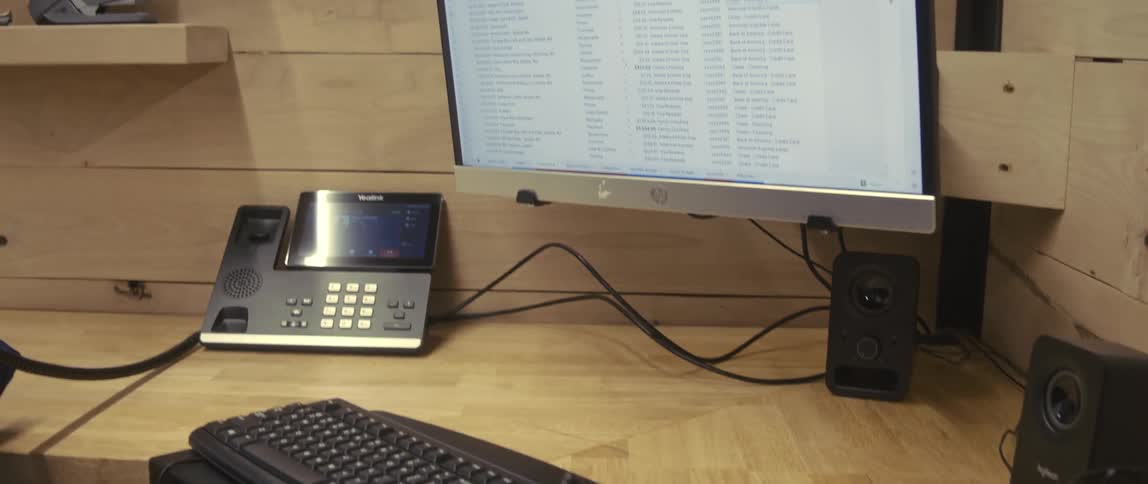 An employee in an office cubicle writes on a pad while on a telephone with email open on a monitor