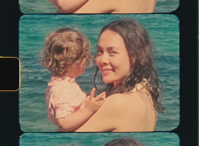 Super 8 film of a mother with her daughter in her arms. They are near the water and the woman is looking back and smiling