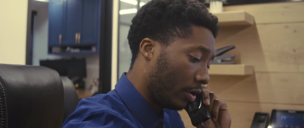 A young man in an office cubicle talking on the phone then leaning forward to take notes