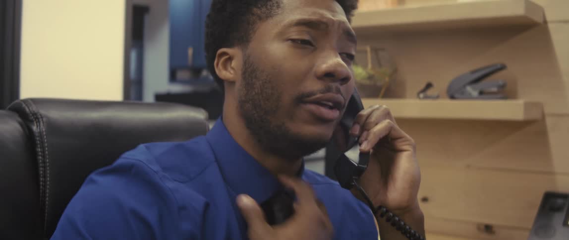A concerned office worker wearing a blue shirt talking on the phone on his cubicle