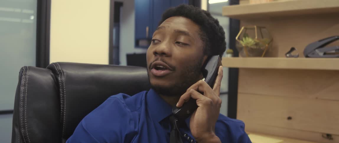 A businessman dressed in a blue collared shirt and a black tie is sitting in a black leather office chair speaking on a phone with a focused expression
