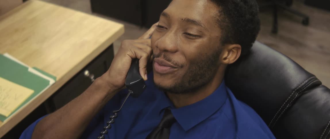 A young man sitting in an office chair and he is talking on the phone and he is smiling
