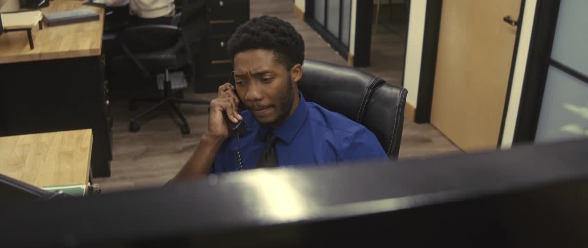 A man in blue formal wear is sitting on an office chair and talking on a phone and he is happy