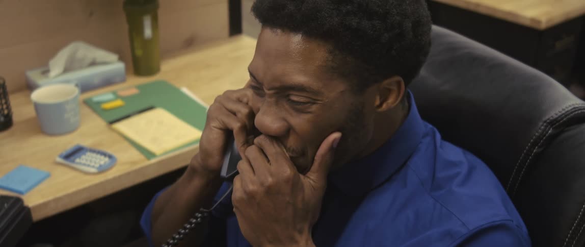 A young man in a blue shirt is sitting in an office talking on the phone and he is very happy