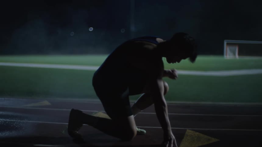 Man squatting and getting into starting position on a running track at night