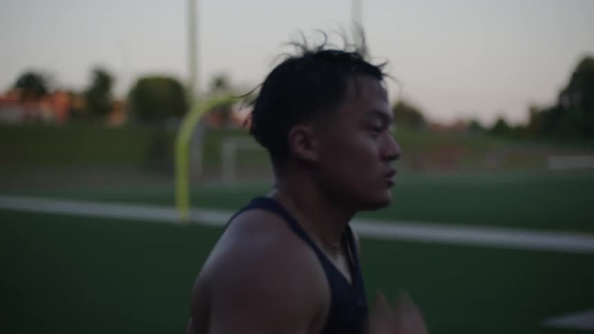 An asian male sweats as he runs around a football field on a running track