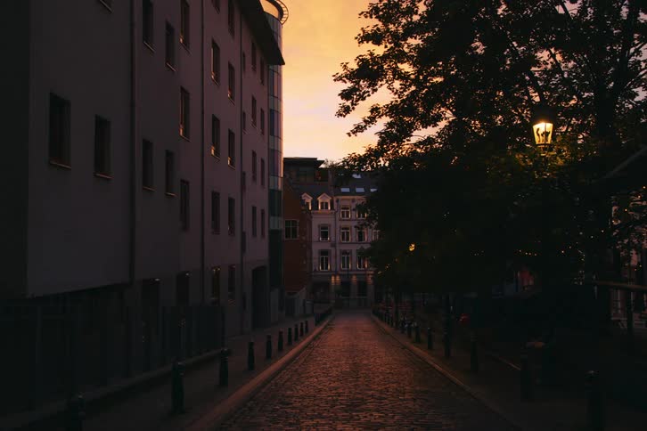 A narrow cobblestone street lined with old buildings illuminated by a single lamppost against a dramatic sunset
