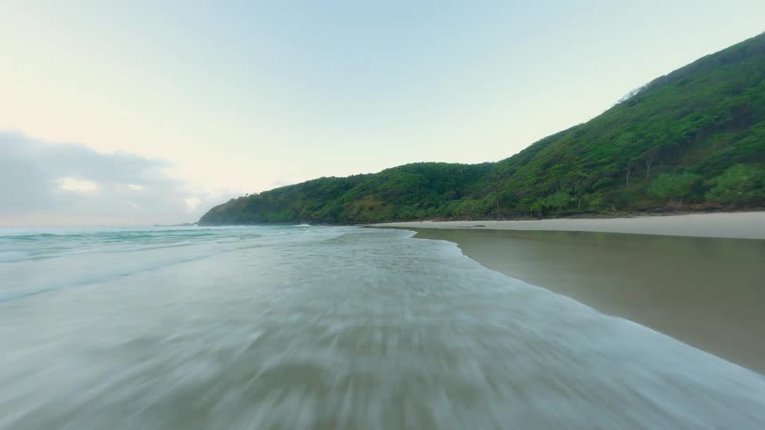 An aerial view of a beach on a coast with sand and stone and there is a forest on the hills around