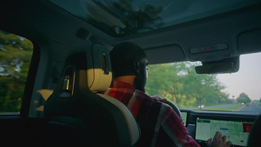 A black man fiddles with a vehicles dashboard computer while driving down a highway
