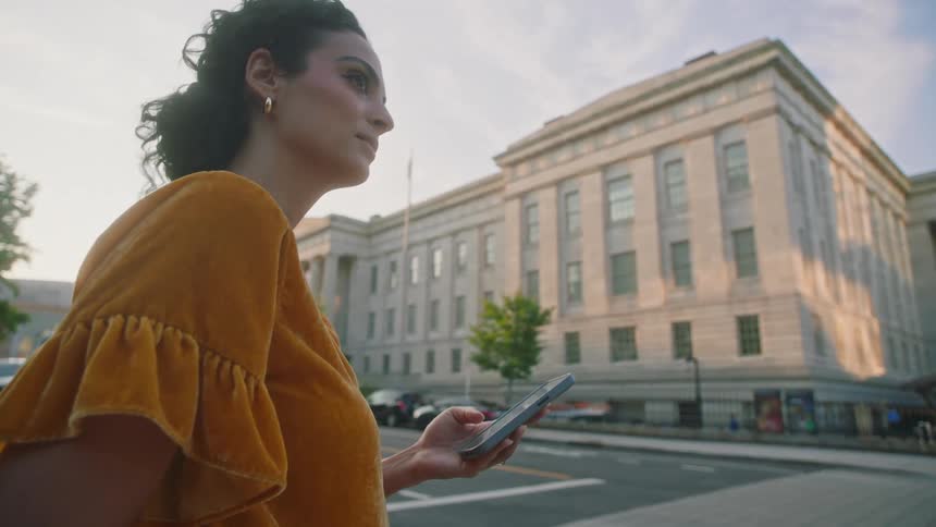 A woman in an orange shirt is standing looking at her cell phone which is in her hand and then she looks forward