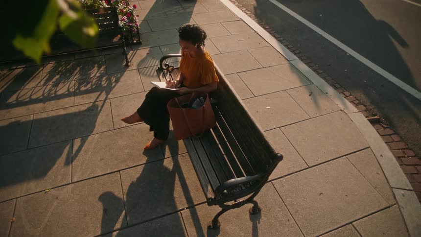 A woman wearing an orange blouse and is sitting on a public bench on a sidewalk and she is writing on a paper