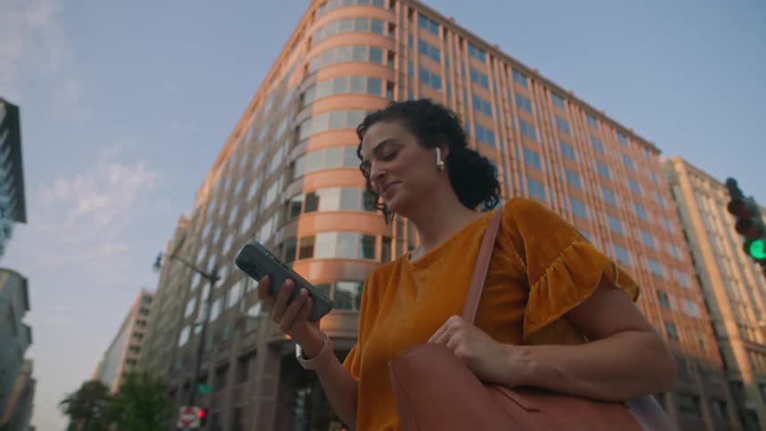 A woman in a yellow shirt and holding a bag on her shoulder and is looking at her cell phone which is in her hand while walking forward