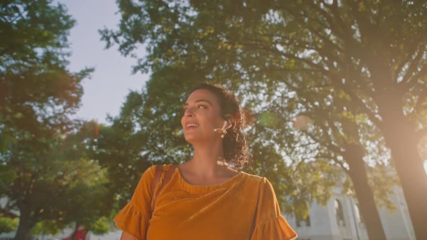 A woman is wearing an orange shirt and she is walking happily in an outdoor environment with trees on the sides