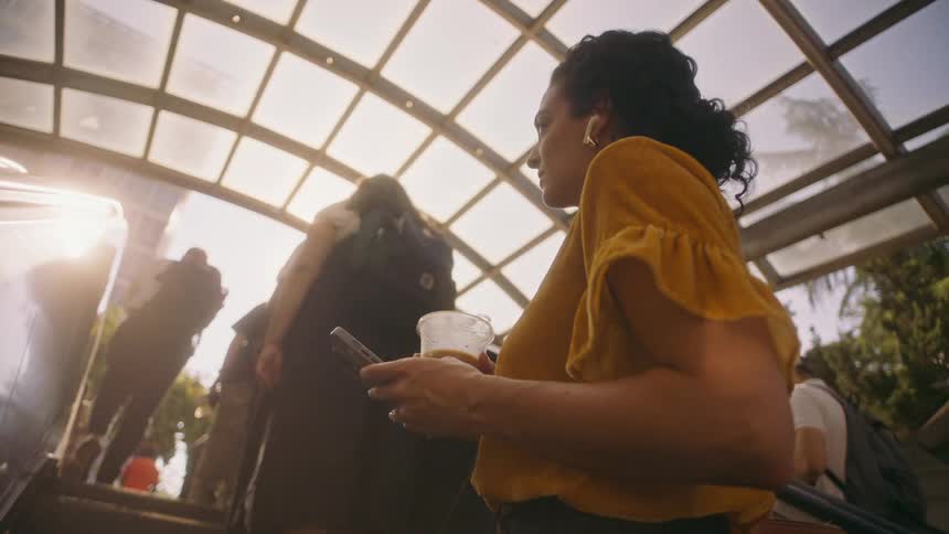A young woman about to come out of an escalator into the street carrying a smart phone and a latte cup while wearing earbuds