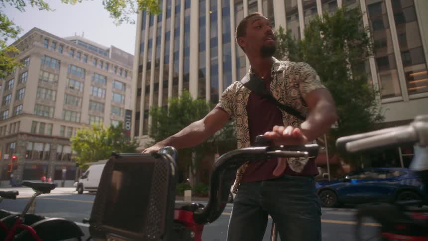A man picks up a bicycle that is parked and he reverses it and starts riding it on an asphalt street and he is happy