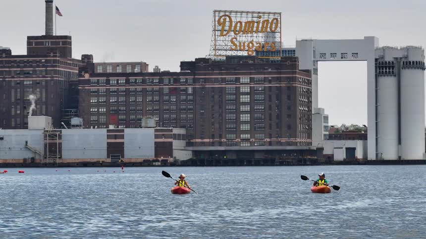 Two kayakers paddle through the water passing a factory in unison