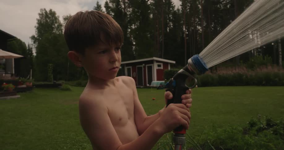 A shirtless boy watering large green plants with a hose in the green backyard