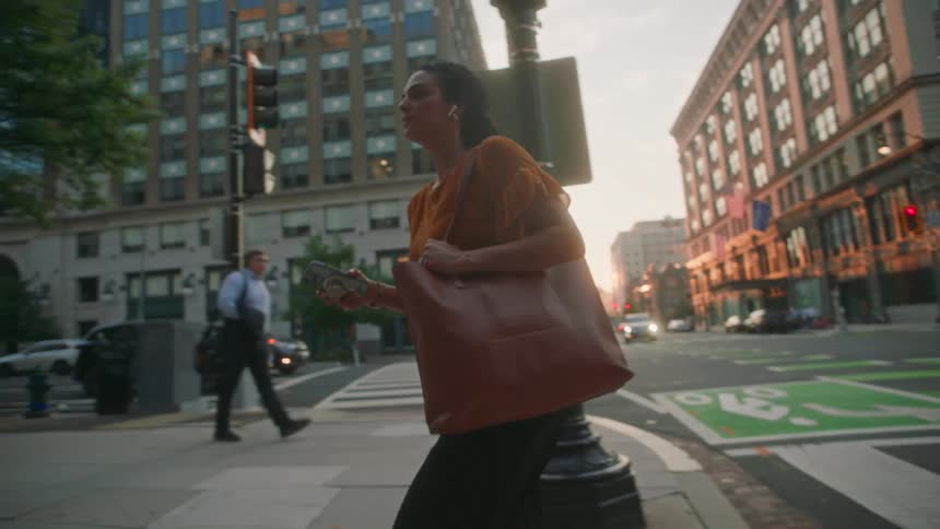 A woman walks down a city street carrying a large brown leather tote bag and a smartphone