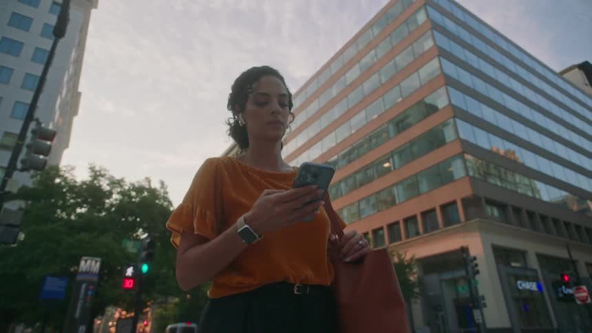 A woman with an orange shirt and is holding a cell phone while walking and behind her there are some commercial buildings