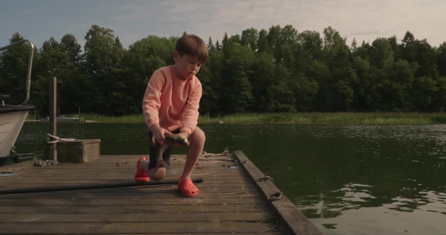 A child stands on a wooden dock throwing a live fish into a lake