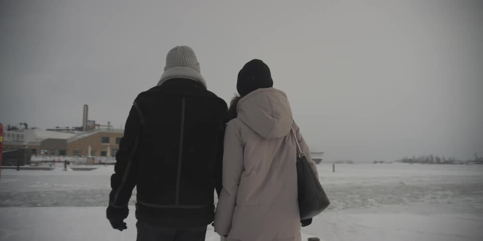 A couple walk hand in hand through a snowy landscape with a city in the background