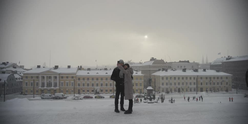 A man and a woman hug and stand in a snowy city as the woman films on a smartphone