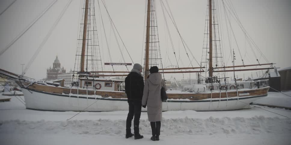 A couple hold hands looking at a sailboat as it snows