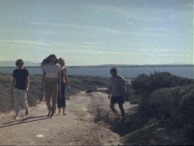 Group of people walk along a dirt trail in a natural setting with water and hills in the background