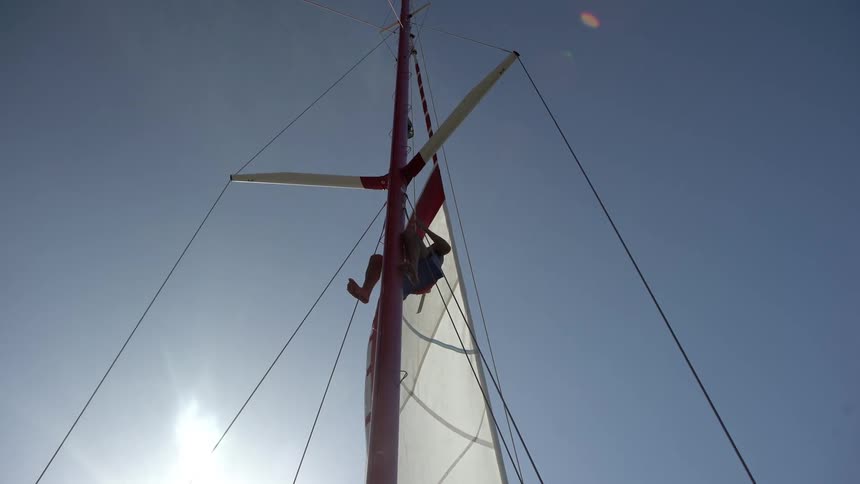 A person climbs the mast of a sailboat with the sun in the background
