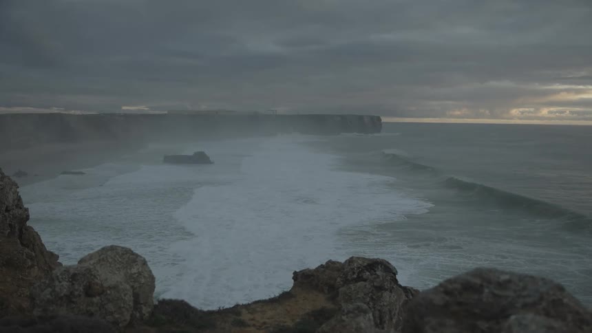 Large ocean waves pound a beach ringed by mountainous terrain