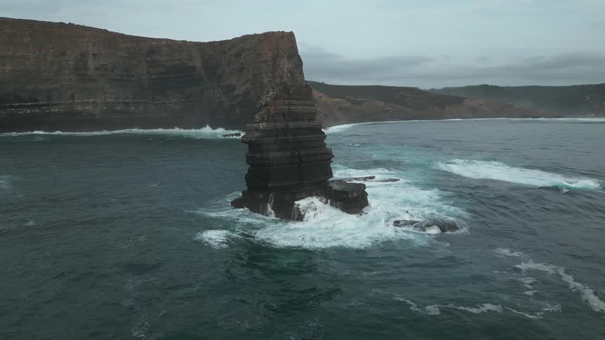 A large layered rock formation stands in the ocean with waves crashing at its base while towering cliffs line the background along the coast