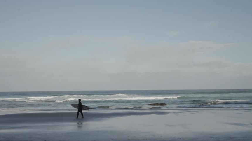 A person holding a surfboard walks on the shore of a beach with waves in the background