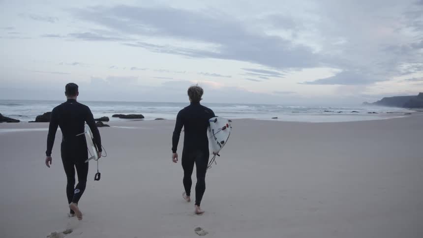 Two men wearing wetsuits walk towards the sea with surfboards on a quiet beach
