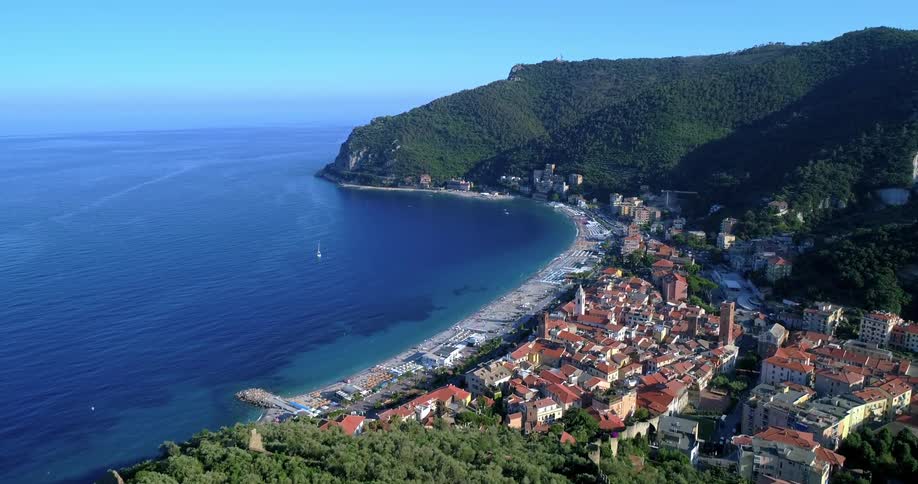 An aerial view of a coastal town with a prominent tower structure nestled between lush green hills and the expansive blue sea