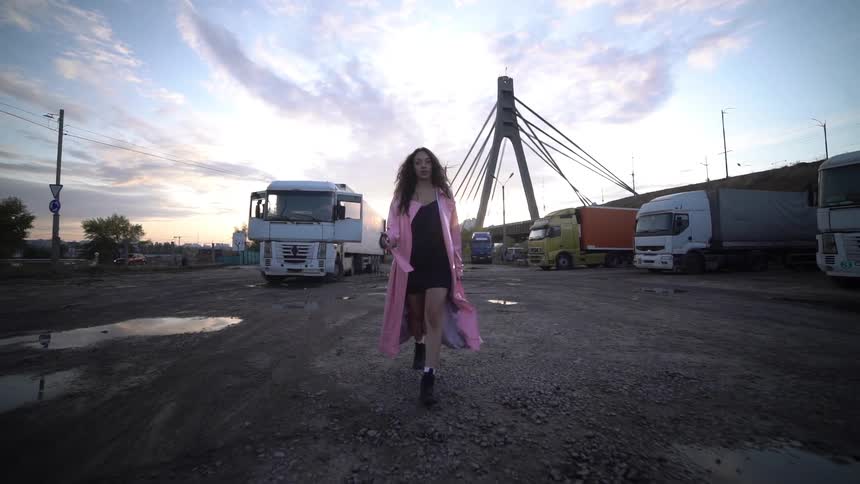 A woman wearing a pink coat walking in the center of a parking area surrounded by trucks during sunrise or sunset