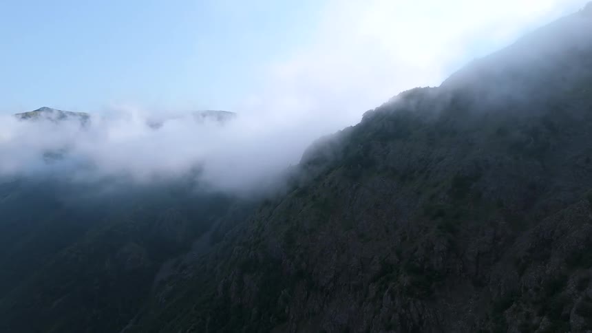 A rocky mountainside is partially shrouded in mist with clouds drifting over the peaks and a clear blue sky above