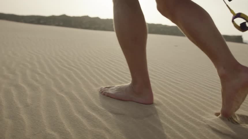 A man in red shorts and a white shirt walks barefoot in the sand holding a white surfboard leaving footprints behind with the ocean visible in the distance