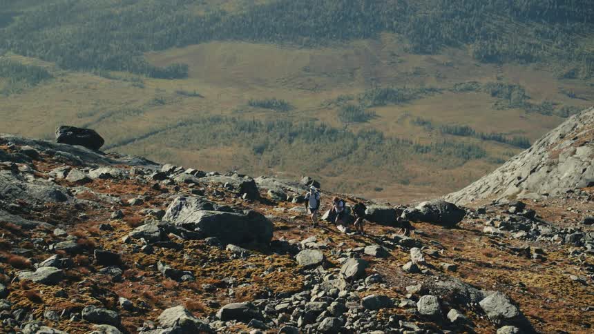 Backpackers pass boulders while hiking up a slope overlooking a forest covered plain on a foggy day