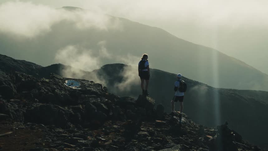 Two hikers stand on top of a mountain and look at the clouds